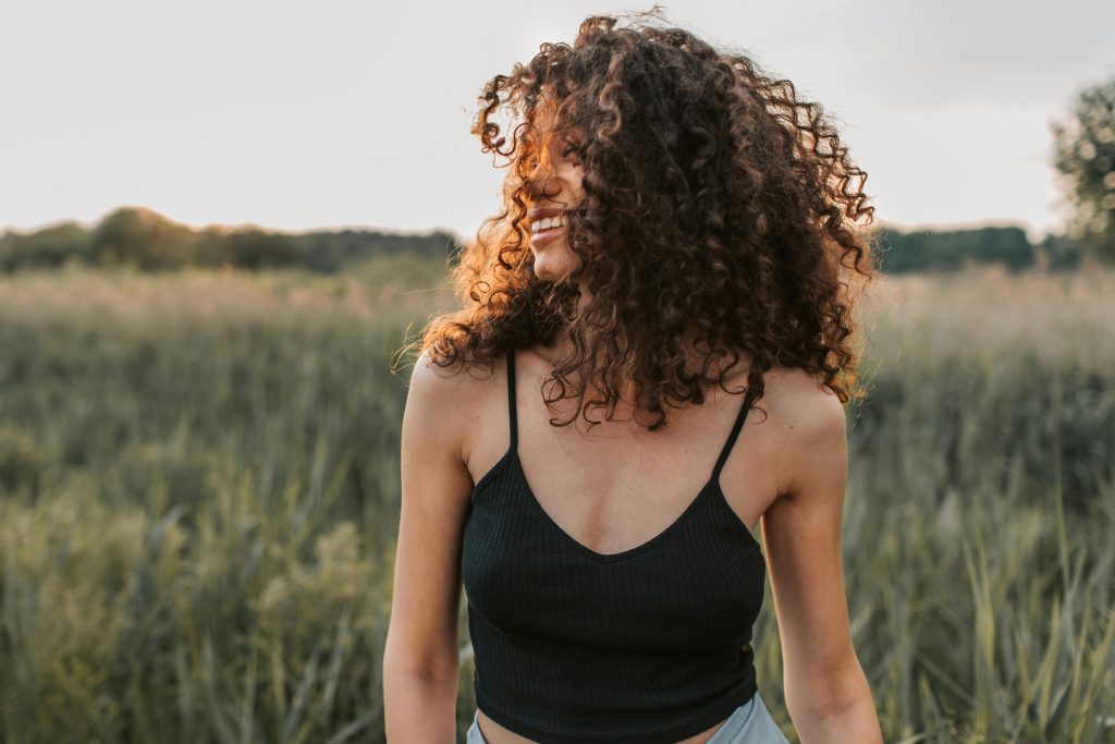 A cheerful woman in casual attire enjoying a sunny day outdoors with natural curly hair.